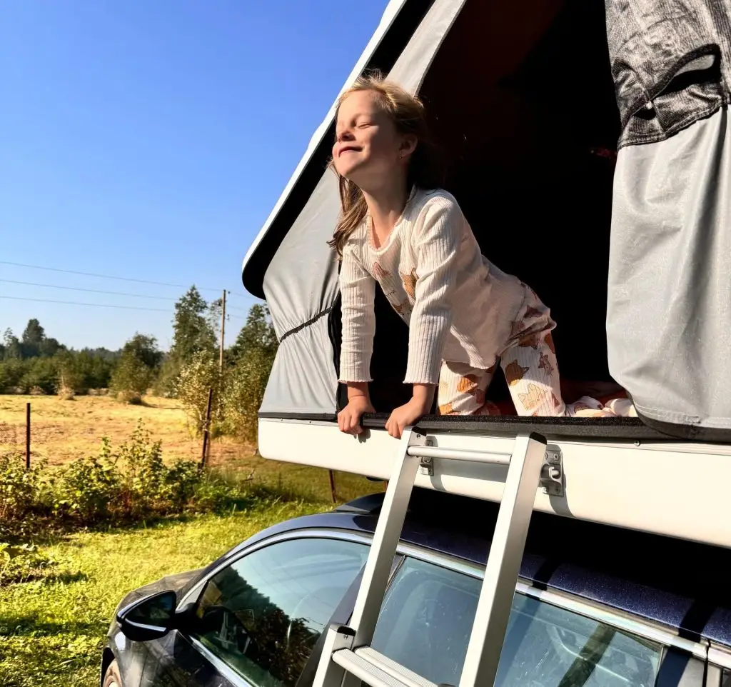 Mazā meitene priecīgi izbauda rītu, izlienot no auto jumta telts